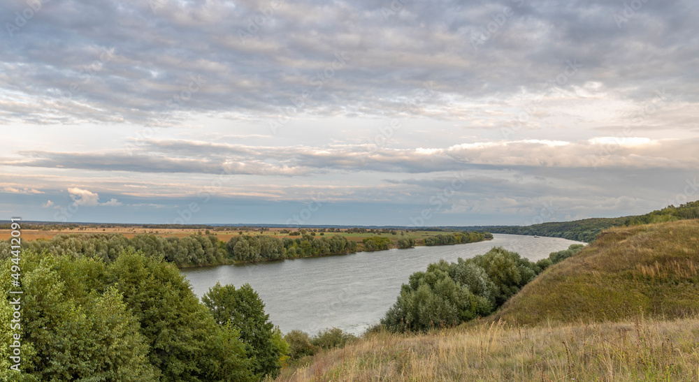 Scenic panoramic view. Green hills, fields and meadows. Sunny summer landscape with a river. Dramatic sky over the horizon. Calm. Clouds over the river.