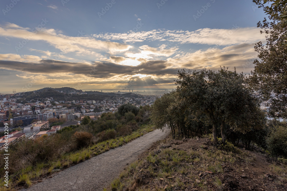 Obraz premium Picturesque landscape of Barcelona from the hill in the early morning. Road in the foreground. Sunbeams through the clouds. Dramatic sky over the city. Autumn in Barcelona, Spain.