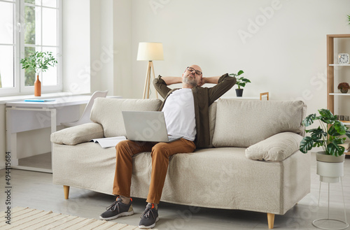 Adult man takes break while working remotely in his home office. Relaxed mature man sitting with hands behind head and laptop computer on his lap on comfortable light beige sofa in modern interior