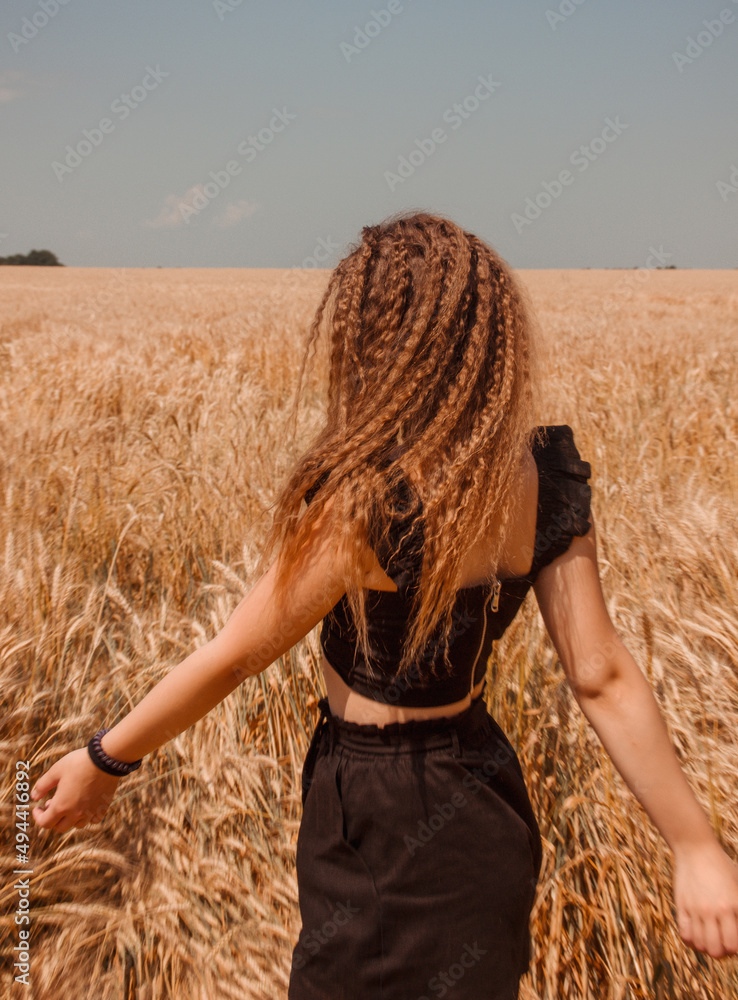 young teen girl with curly hair in black summer costume is standing from the back and hugs the golden wheat field with blue sky background at sunny day. lifestyle concept, free space