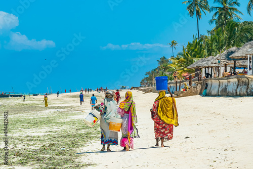 Zanzibar, Tanzania - December 19, 2021: Local women walk by the white sand beach, Zanzibar, Tanzania