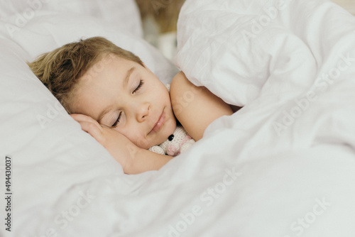 Beautiful blond toddler child, boy, sleeping in bed with teddy bear toy