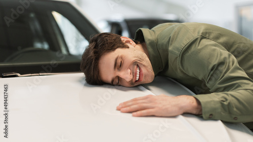 Happy young Caucasian guy hugging his brand new car, buying vehicle at dealership centre, panorama with copy space