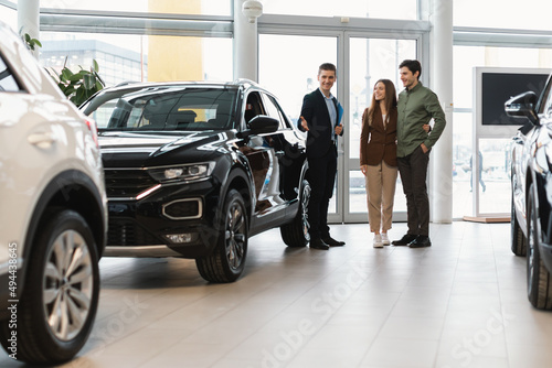 Confident salesman showing new car to young Caucasian couple at auto dealership