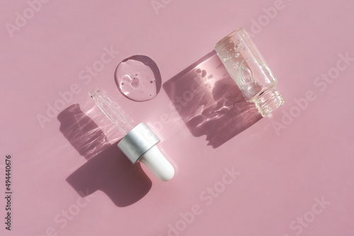A bottle of cosmetic gel and a glass pipette cast shadows on a pink background. Flat lay.