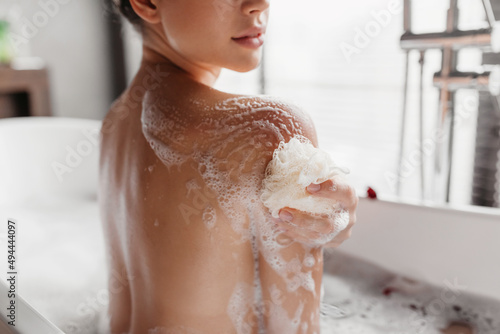 Home spa, hygiene concept. Young lady taking bath and rubbing her skin with sponge, relaxing in bathroom, closeup