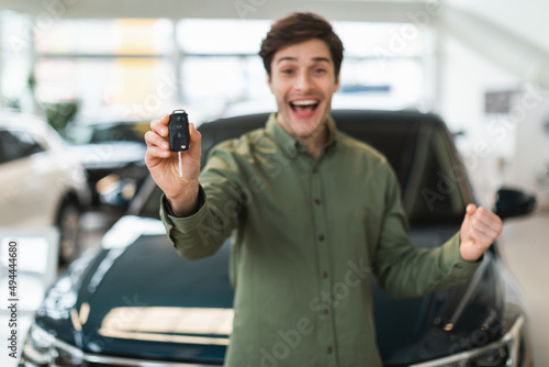 Excited young Caucasian man showing new car key at camera, gesturing YES, celebrating purchase of auto at dealership