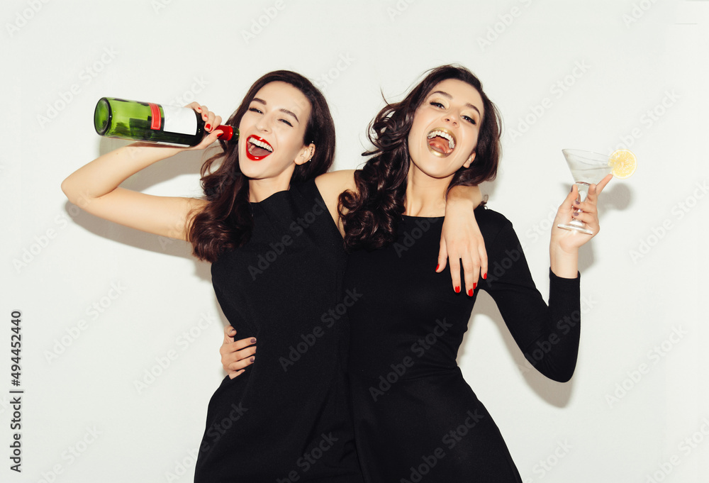 Two brunette sisters with long hair drink an alcoholic cocktail and wine. Festive girls in evening dress laugh and smile. A bottle of wine. White background. Two brunette sisters with long hair drink an alcoholic cocktail and wine. Festive girls in evening dress laugh and smile. A bottle of wine. White background.