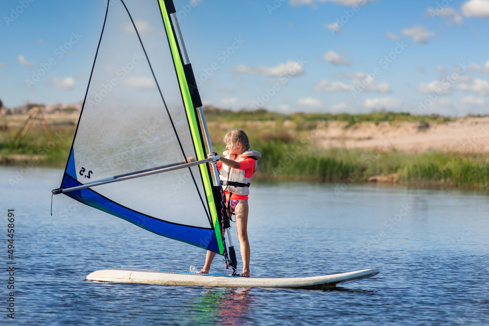 Cute little caucasian blond child girl enjoy having fun learning to surf on winsurfing board at freshwater pond lake or river hot sunny day. Summer outdoors child healthy sport recreation activities