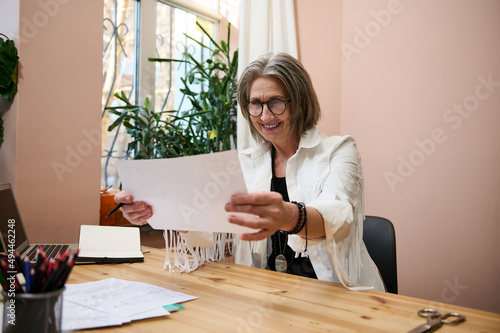 Inspired mature business woman female fashion designer looks at drawn sketches for clothes while sitting at wooden table in her design studio office or workshop and smiling with cheerful toothy smile