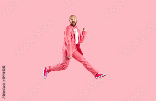 Happy excited funny young man in suit and sneakers having fun jumping on pink background. Cheerful Caucasian bald bearded man in pink formal suit is having fun and jumping. Banner. Full length.