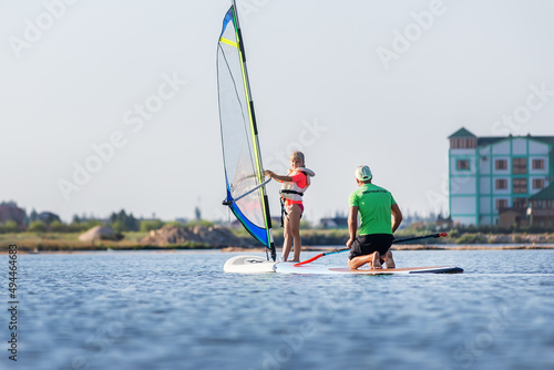 Cute little caucasian blond child girl enjoy having fun learning to surf on winsurfing board at freshwater pond lake or river hot sunny day. Summer outdoors child healthy sport recreation activities