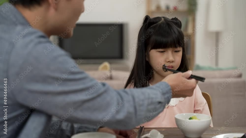 selective focus of unhappy Asian daughter turning head while refusing food his father is feeding her during lunch time in dining room at home selective focus of unhappy Asian daughter turning head while refusing food his father is feeding her during lunch time in dining room at home