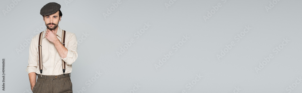 pensive man in shirt with suspenders and retro style hat posing with hand in pocket isolated on grey, banner. pensive man in shirt with suspenders and retro style hat posing with hand in pocket isolated on grey, banner.