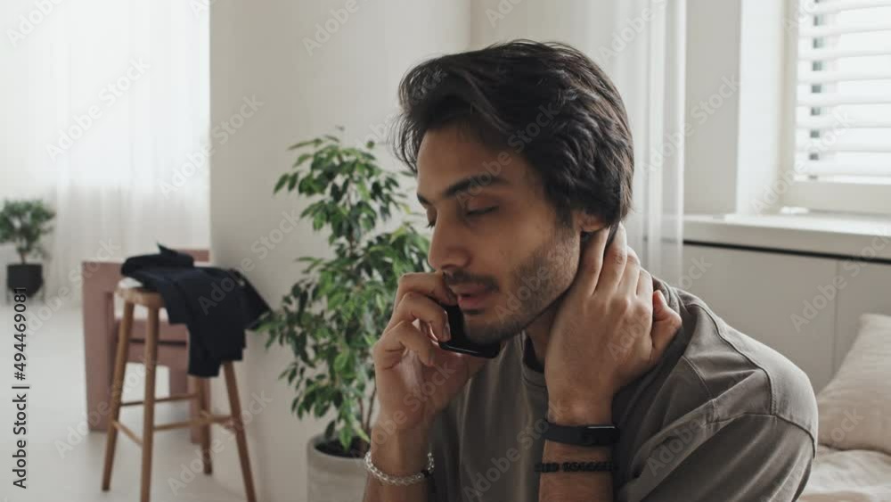 Chest-up of young Middle Eastern man sitting in bedroom in modern apartment, talking on cellphone in morning Chest-up of young Middle Eastern man sitting in bedroom in modern apartment, talking on cellphone in morning