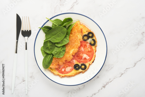 Omelette with tomatoes, black olives, cottage cheese and green spinach herbs on white plate on grey concrete background. Delicious breakfast. Healthy breakfast food. Top view.