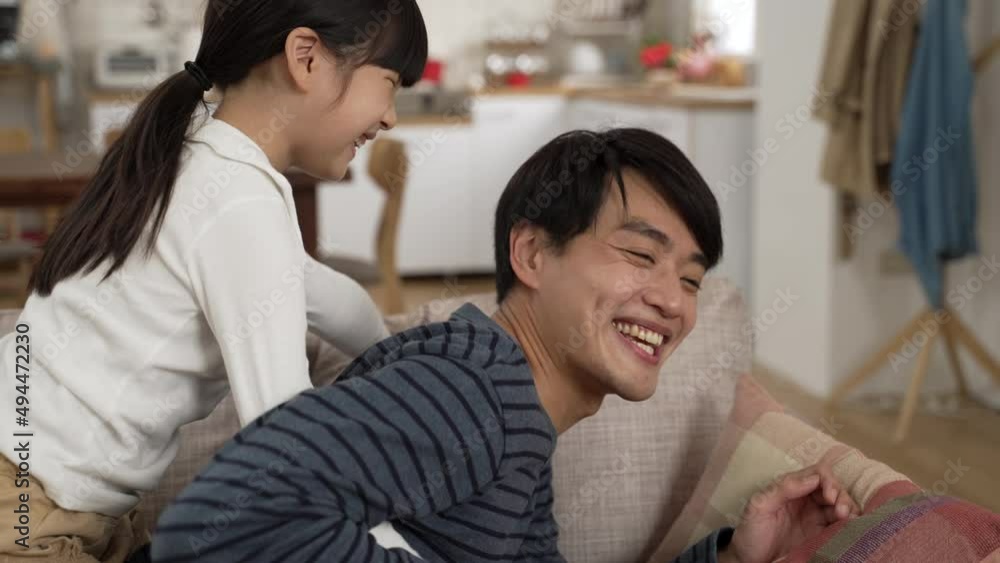 cute asian girl hugging and kissing her father’s face from behind on the sofa while he is lying relaxing. she sits and plays on the smiling man’s back in the living room at home cute asian girl hugging and kissing her father’s face from behind on the sofa while he is lying relaxing. she sits and plays on the smiling man’s back in the living room at home