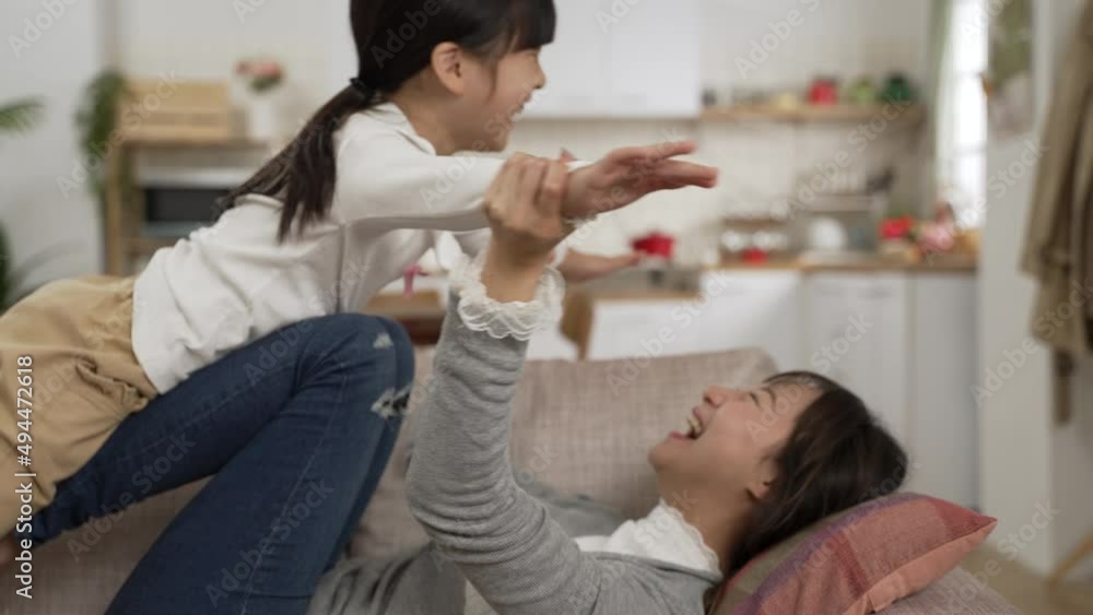 Side view Cheerful Asian mother lying on sofa and holding up daughter on legs at home. The girl open arms like she’s flying Side view Cheerful Asian mother lying on sofa and holding up daughter on legs at home. The girl open arms like she’s flying