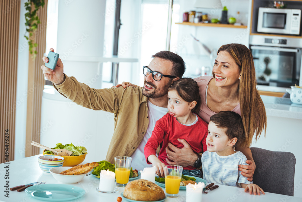 Smiling young family with small preschool children sitting at a table in the kitchen, taking a self-portrait on the phone together, happy parents with small children having fun and taking selfies