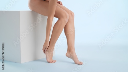 Slim woman strokes her feet sitting on the white cube podium on pale blue background | After woman shaving concept