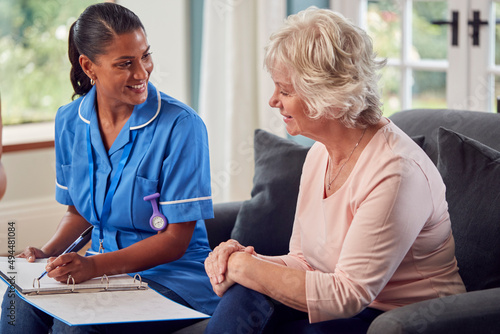 Senior Woman At Home Talking To Female Nurse Or Care Worker In Uniform Making Notes In Folder