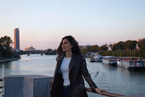 Young and beautiful woman with dark and curly hair is sightseeing in Seville. In the background the river guadalquivir and part of the city in the golden hour. Tourism and holidays concept.