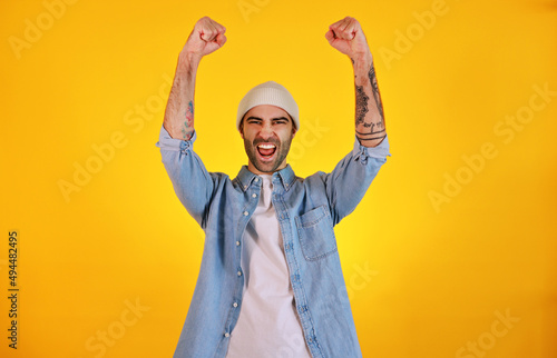 Winner. Rise up hands. Studio shot of smiliing handsome man in jeans and white hat on yellow background. Tatoo and beard. Emotional concept. photo