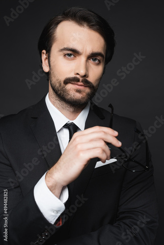 businessman in suit holding glasses and looking at camera isolated on dark grey.