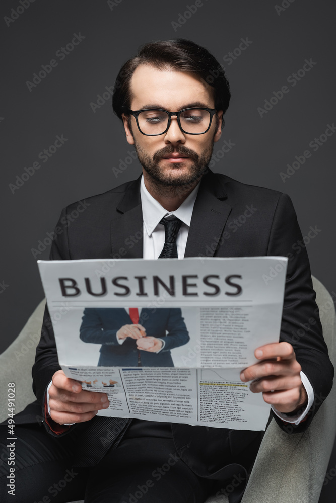businessman in suit and glasses sitting on armchair and reading business newspaper on dark grey. businessman in suit and glasses sitting on armchair and reading business newspaper on dark grey.