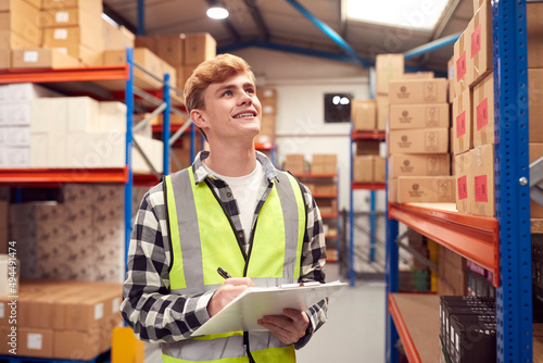 Foto Male Intern Working Inside Warehouse Checking Stock On Shelves Using Clipboard