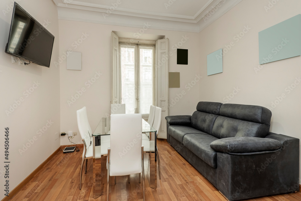 Living room with a three-seater black leather sofa and a glass dining table with white leather chairs and a flat-screen TV on the wall Living room with a three-seater black leather sofa and a glass dining table with white leather chairs and a flat-screen TV on the wall