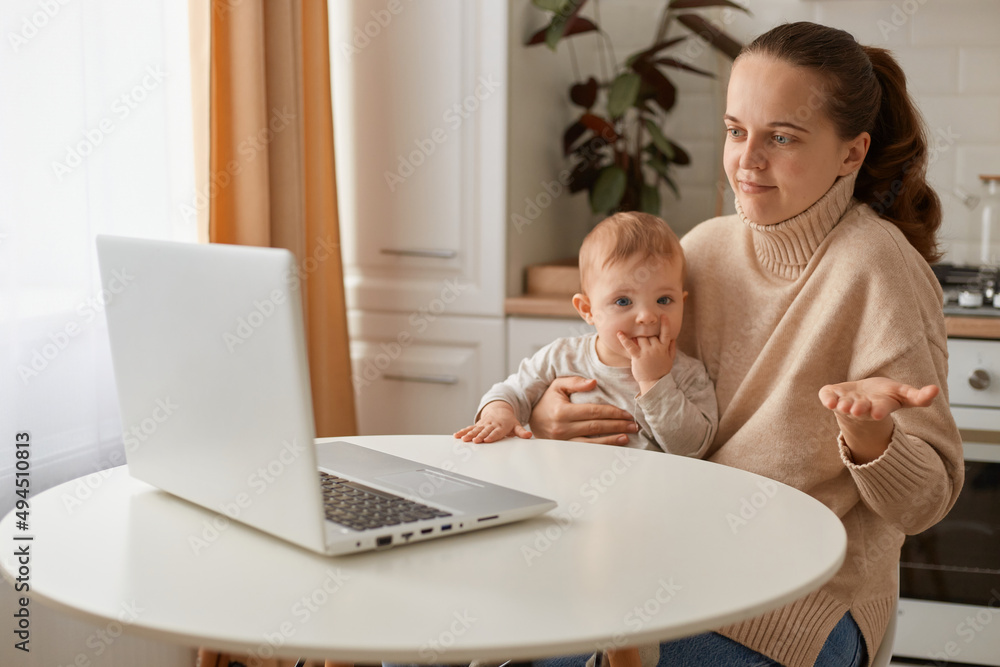 Confused woman with dark hair wearing beige sweater sitting in kitchen and working on laptop and holding infant daughter, shrugging and spreading hands, looks doubtful. Confused woman with dark hair wearing beige sweater sitting in kitchen and working on laptop and holding infant daughter, shrugging and spreading hands, looks doubtful.