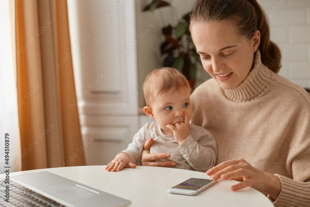 Smiling joyful woman with dark hair wearing beige sweater sitting in kitchen and using mobile phone, expressing positive emotions, spending time with infant daughter. Smiling joyful woman with dark hair wearing beige sweater sitting in kitchen and using mobile phone, expressing positive emotions, spending time with infant daughter.