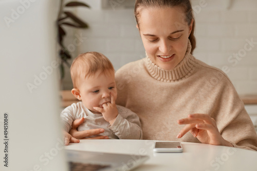 Portrait of adorable woman with dark hair wearing beige sweater sitting in kitchen and working on laptop, using cell phone and holding infant baby in hands.