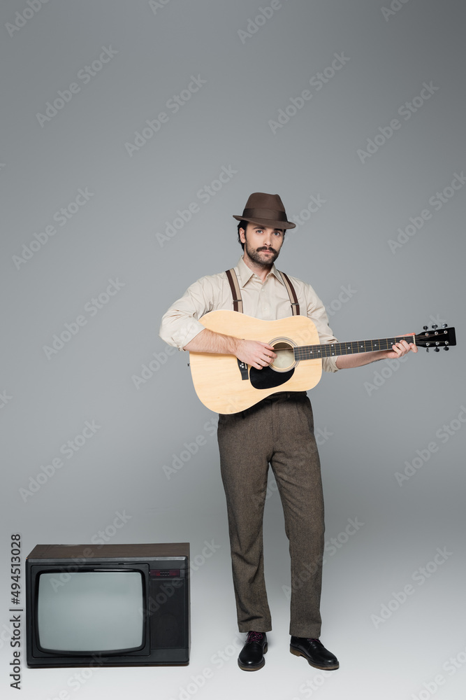 full length of man retro style clothing playing acoustic guitar and standing near antique tv on grey. full length of man retro style clothing playing acoustic guitar and standing near antique tv on grey.