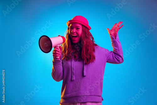 Portrait of young emotive girl talking on megaphone isolated over blue studio background in pink neon light photo