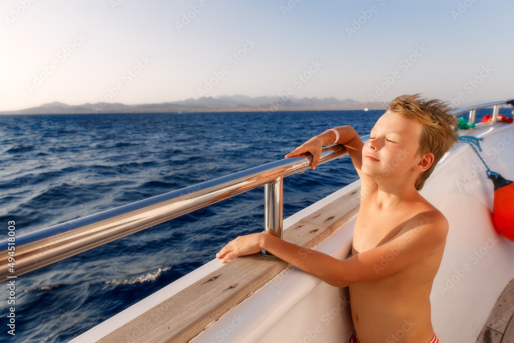 Young boy standing on a deck of yacht closing eyes and enjoying the trip in sunny day. Kid is wearing shorts 