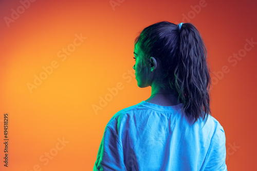Back view portrait of young girl with ponytail in white T-shirt posing isolated over gradient orange background in neon