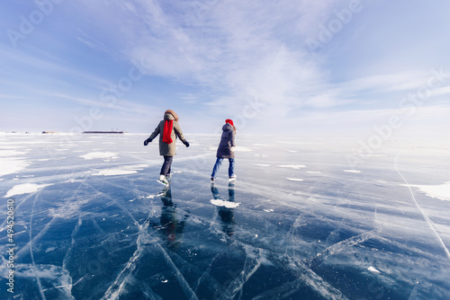 Two young women friends in red cap are skating on ice frozen of Baikal lake, winter sunny day