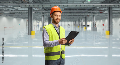 Engineer wearing a reflective vest and helmet holding a clipboard and looking at camera in a warehouse