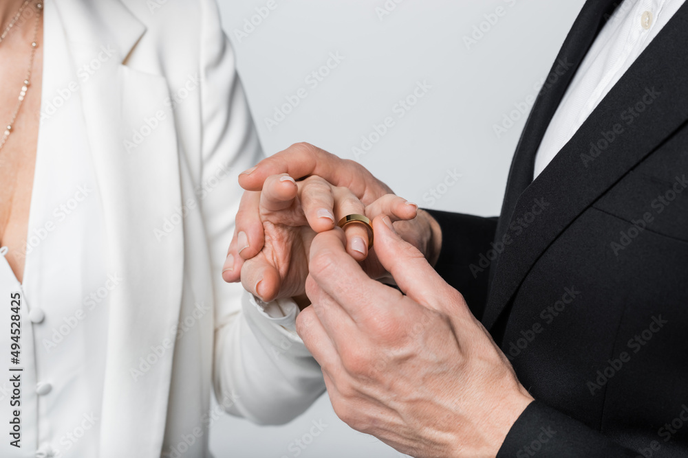 Obraz premium Cropped view of groom wearing ring on finger of bride in formal wear isolated on grey.