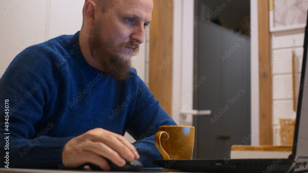 A bald man with a beard sits at a table at home drinking his morning coffee and typing on a smartphone, the beginning of the working day with morning coffee and news from the Internet. A bald man with a beard sits at a table at home drinking his morning coffee and typing on a smartphone, the beginning of the working day with morning coffee and news from the Internet.