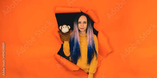 Young woman with soft puppet toy on hand looking out of hole of orange background. Pretty female with puppet monkey. Concept of puppet show. © Anton Dios