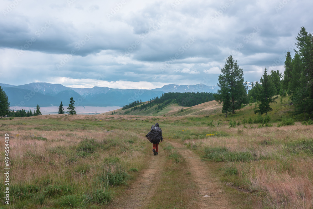 Tourist walks through hills and forest towards bad weather. Hiker on way to large snow mountain range under rainy cloudy sky. Man in raincoat in mountains in overcast. Traveler goes towards adventure.