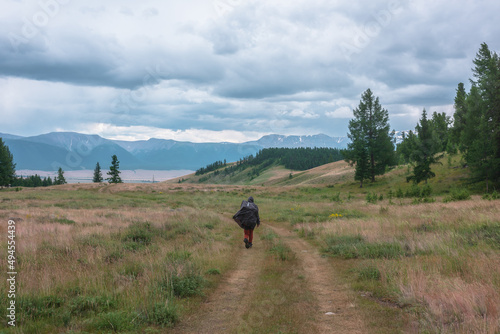 Tourist walks through hills and forest towards bad weather. Hiker on way to large snow mountain range under rainy cloudy sky. Man in raincoat in mountains in overcast. Traveler goes towards adventure.