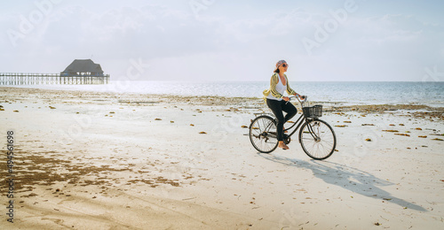 Young woman dressed light summer clothes riding old vintage bicycle with front basket on the lonely low tide ocean white sand coast on Kiwengwa beach on Zanzibar island, Tanzania.
