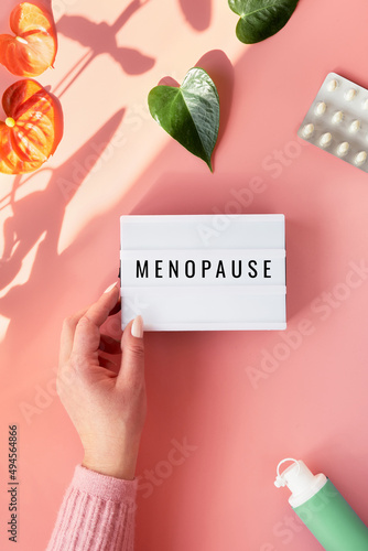 Text Menopause on light box in hand. HRT Replacement hormone therapy, HRT concept. Pink background with exotic leaves, flowers, pills, estrogene gel. Sunlight, long shadows. photo