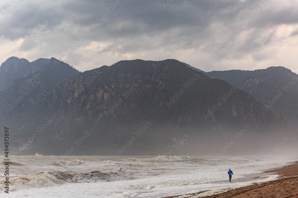 A man walks along the beach on the Mediterranean Sea in Turkey, Antalya, against the backdrop of big waves and mountains, cloudy weather, rain, huge.