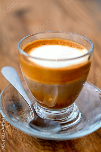 Macchiato coffee drink with foam and layers in glass cup with a spoon on wooden background. Macro cryptography, selective focus, close up and soft focus with bokeh