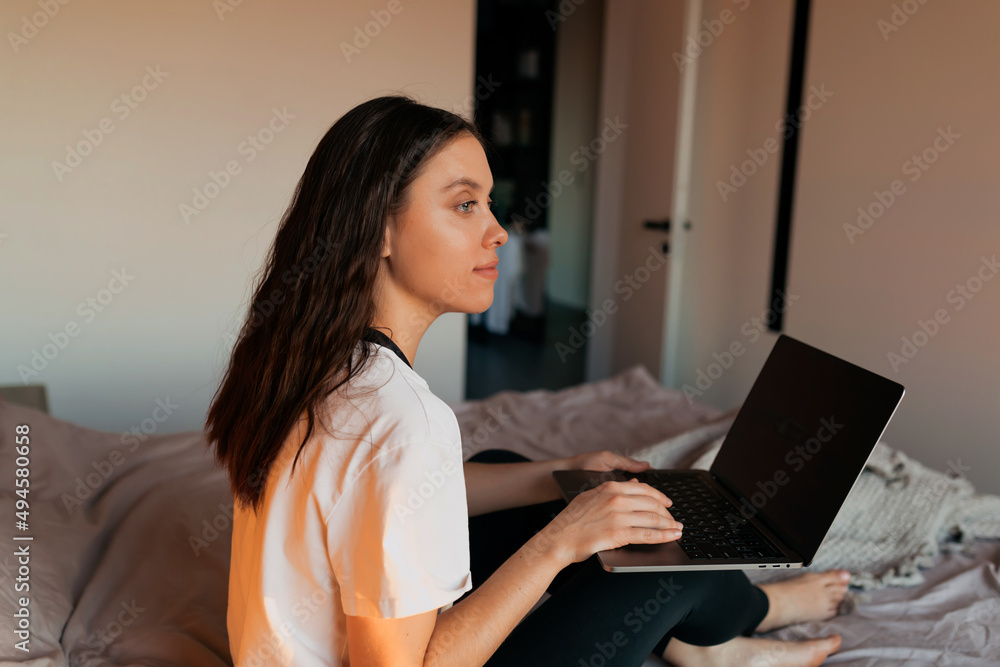 Close up portrait from back of adorable lovely girl is typing on laptop while sitting in the bed after waking up. Charming lady is working on her weekend Close up portrait from back of adorable lovely girl is typing on laptop while sitting in the bed after waking up. Charming lady is working on her weekend